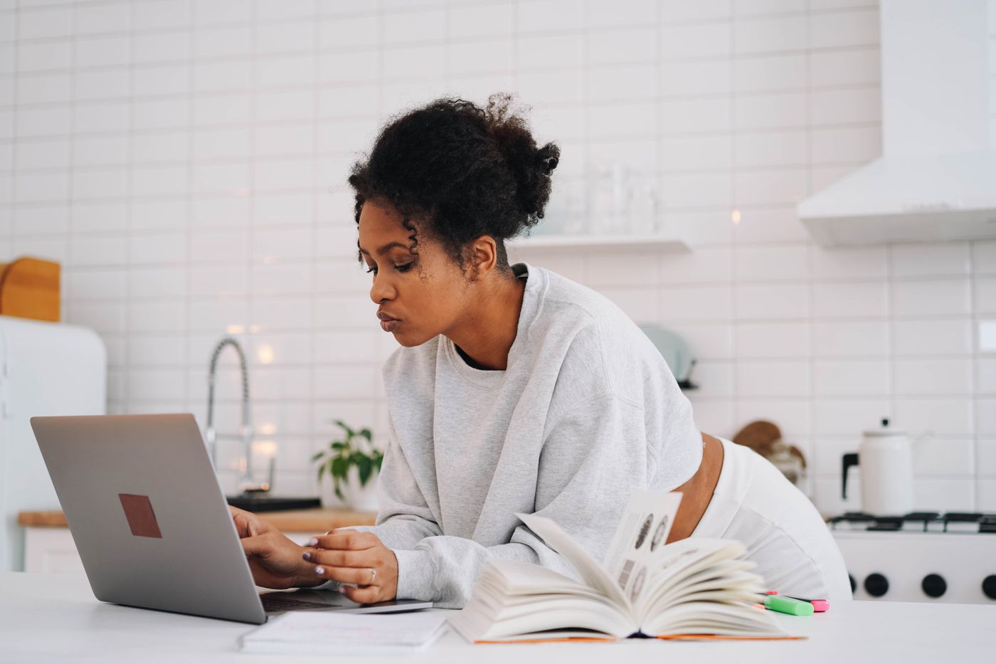 A woman working through an online course on a laptop at a kitchen counter, with an open course book and notes beside her.