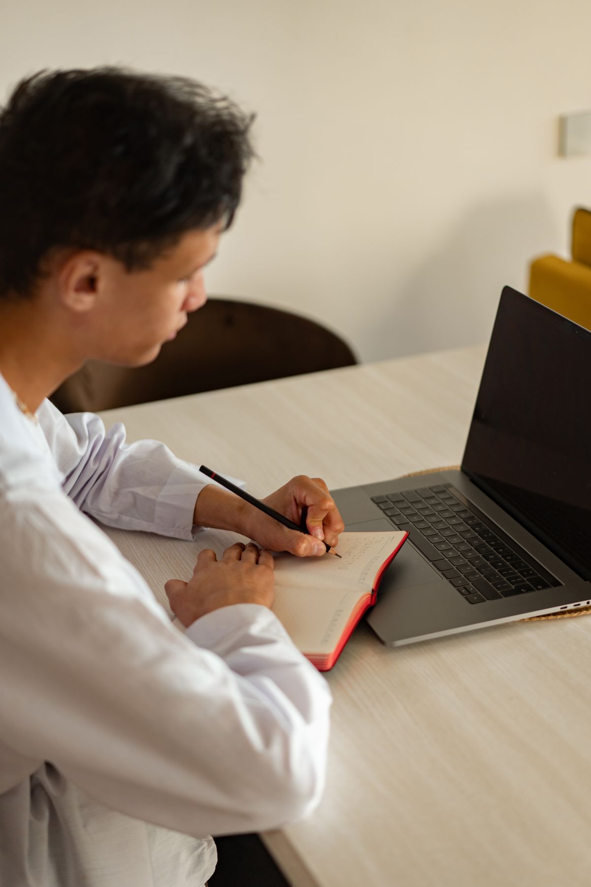 A man taking notes in a notebook beside an open laptop at a dining table.